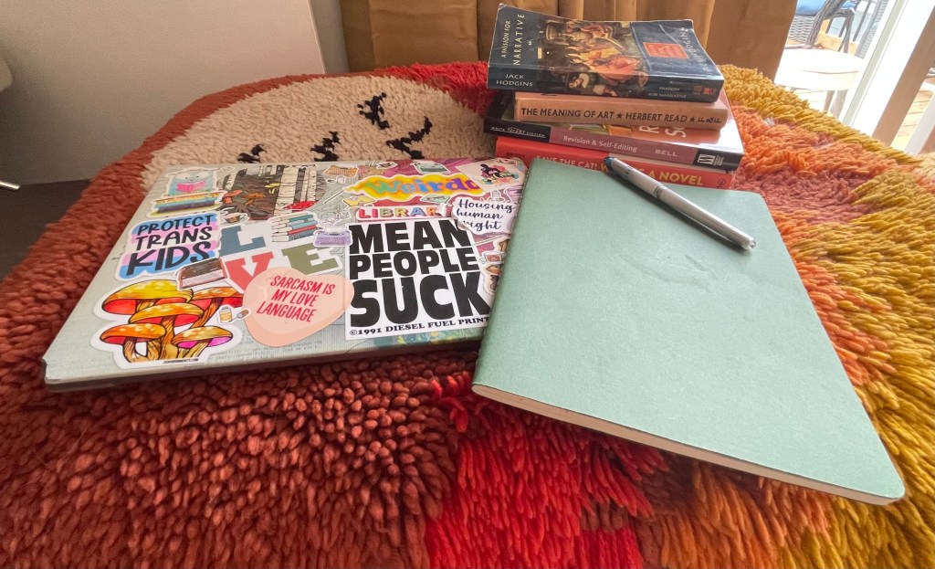 A photo of my laptop covered in stickers, a green notebook with a pen on top, and a stack of books in the top right corner.
